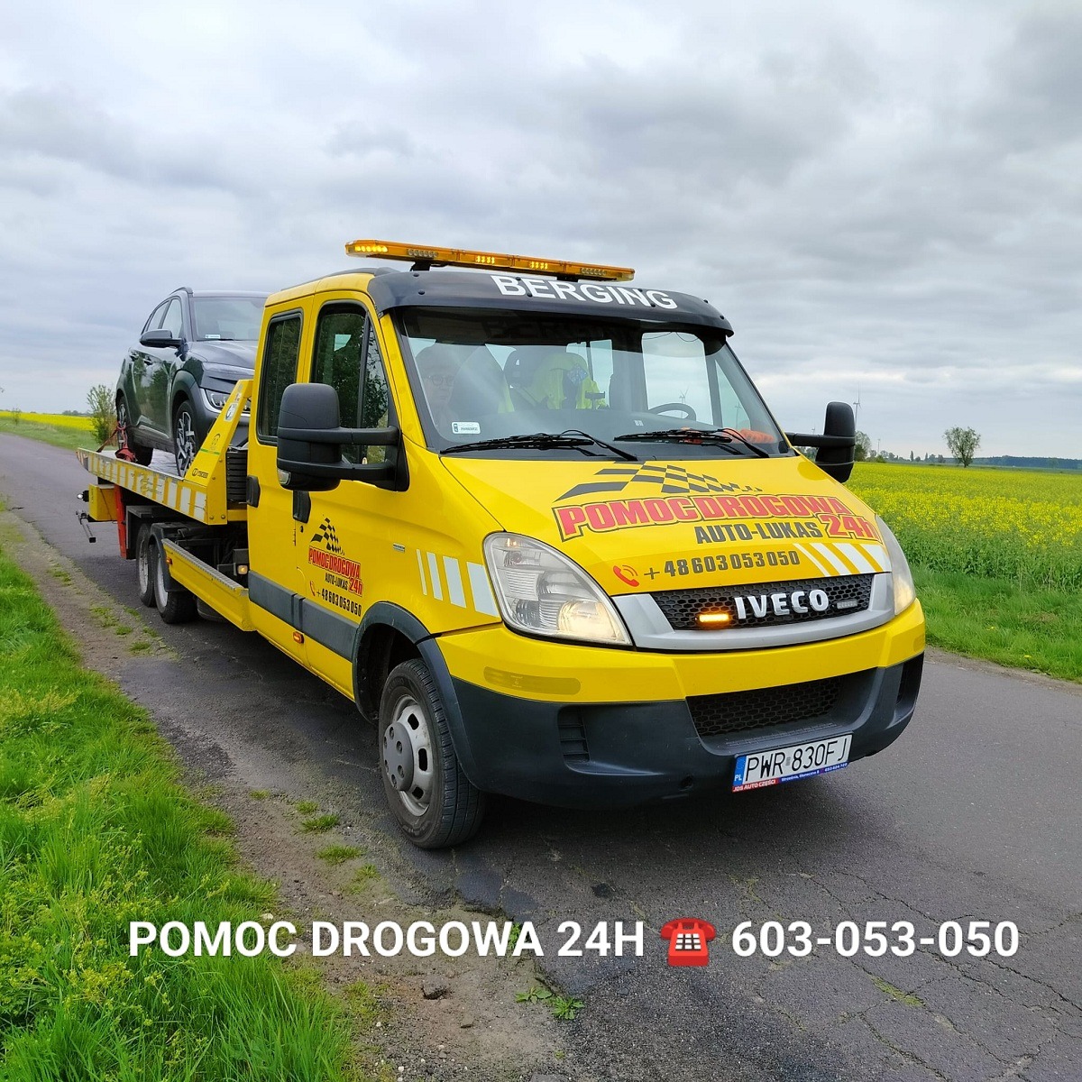 Żółta laweta Iveco z załadowanym szarym samochodem osobowym na drodze asfaltowej z polami rzepaku w tle, oświetlenie ostrzegawcze włączone.