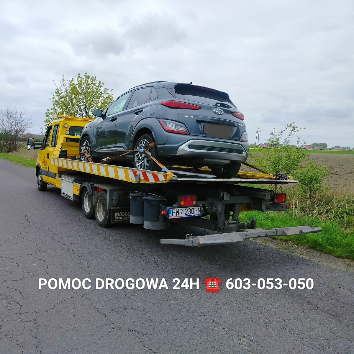 Żółta platforma z szarym Hyundai Kona zabezpieczonym pasami transportowymi, widok z tyłu na drodze wiejskiej, tablica rejestracyjna PWR 230FN.