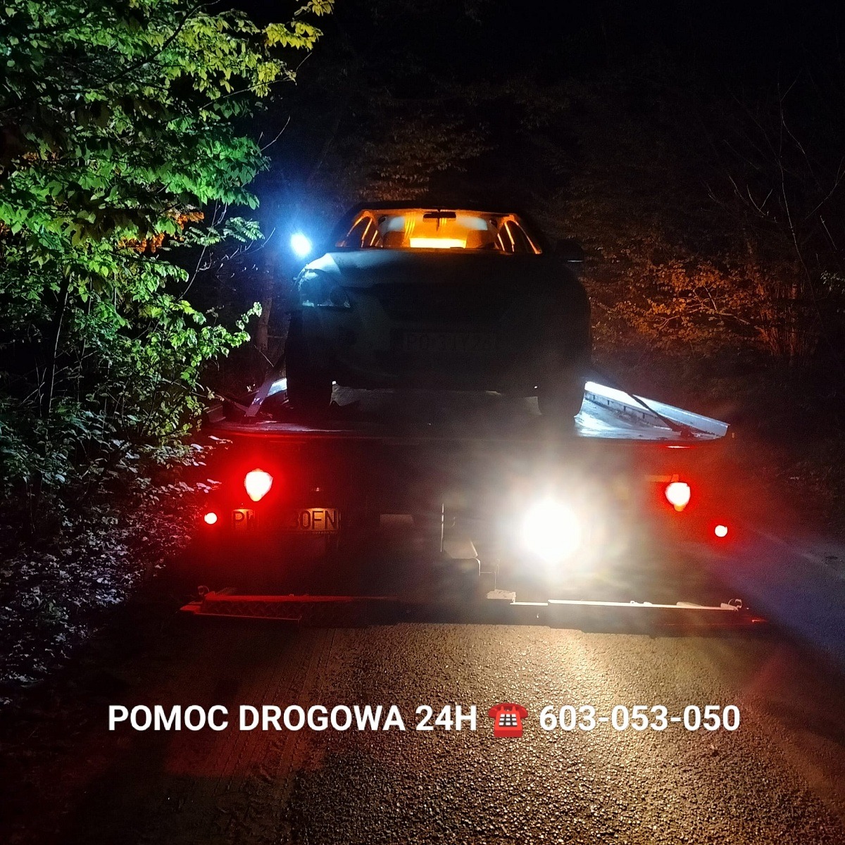 Nocne ujęcie lawety z uszkodzonym samochodem osobowym na platformie, światła awaryjne włączone, widoczne drzewa w tle, na dole kadru napis 'POMOC DROGOWA 24H' z numerem telefonu.