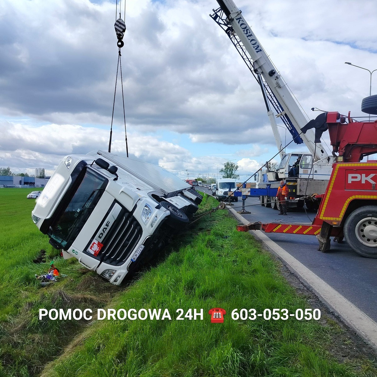 Przewrócona biała ciężarówka DAF na poboczu, podnoszona przez dźwig firmy Koslom, widoczni pracownicy w pomarańczowych kamizelkach, na dole zdjęcia napis POMOC DROGOWA 24H z numerem telefonu.