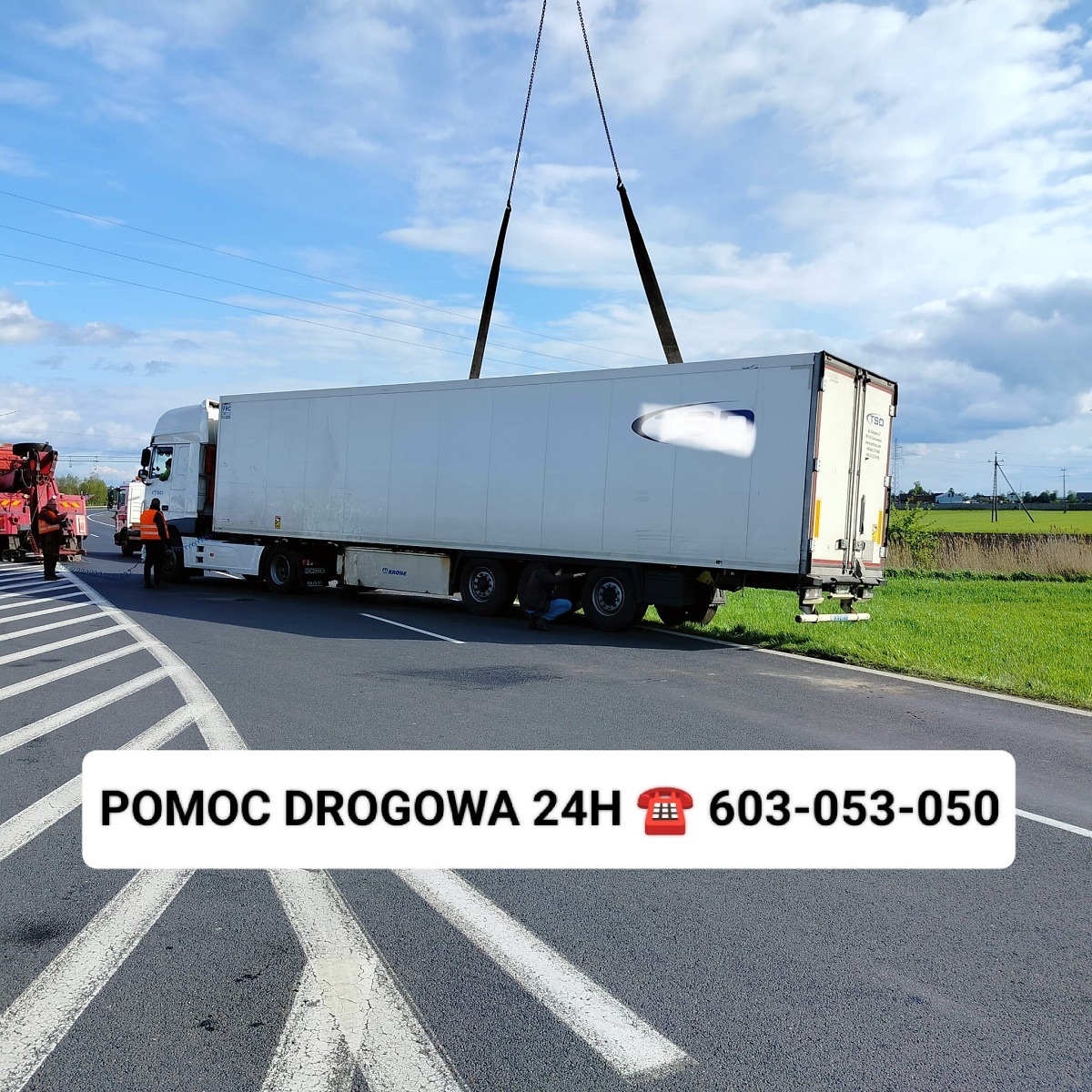 Ciężarówka z naczepą podnoszona przez dźwig na drodze, widoczna pomoc drogowa i osoby w odblaskowych kamizelkach.