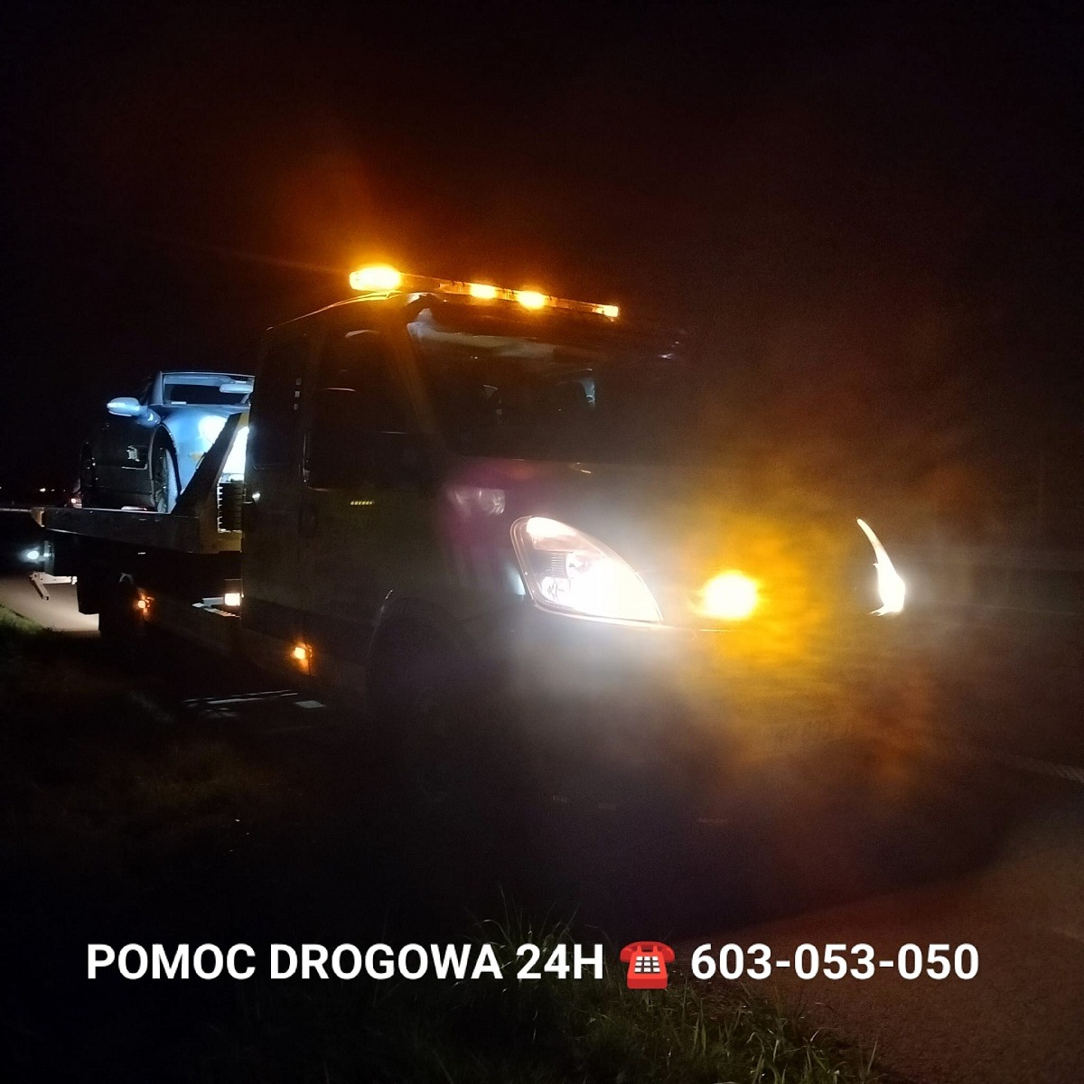 Nocne ujęcie lawety z niebieskim samochodem osobowym na platformie, oświetlonej światłami ostrzegawczymi i reflektorami, w tle ciemność i pobocze drogi, u dołu widoczny napis 'POMOC DROGOWA 24H'...