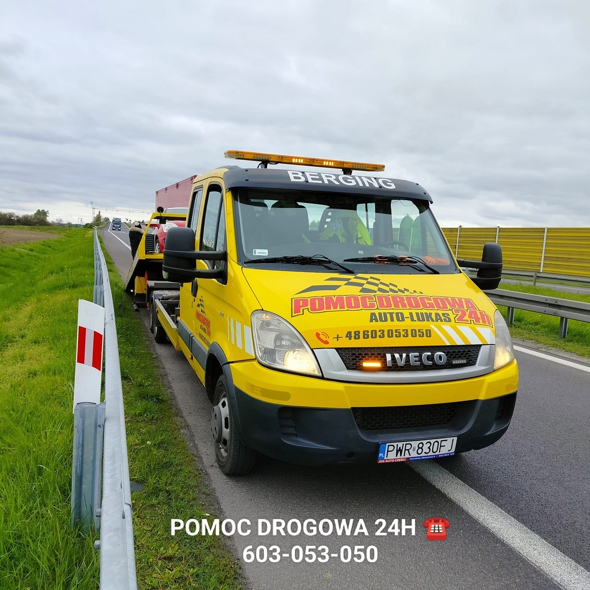 Żółta laweta marki Iveco na poboczu autostrady, z załadowanym czerwonym samochodem osobowym, widoczne oznaczenia firmy 'POMOC DROGOWA AUTO-LUKAS 24h' oraz numer telefonu, pochmurne niebo w tle.