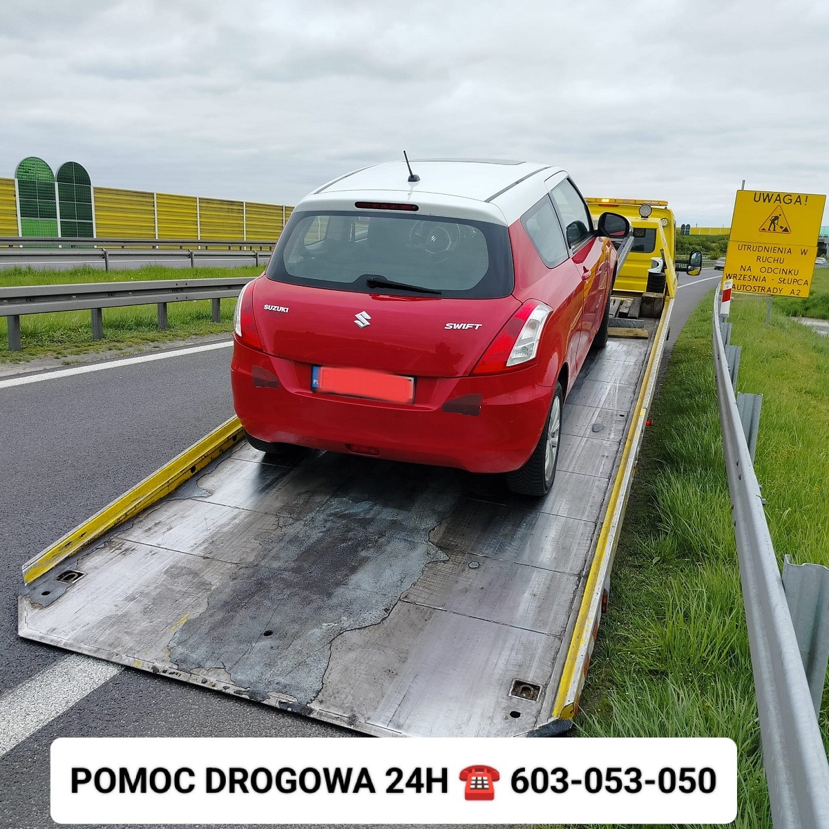 Czerwony Suzuki Swift z białym dachem na platformie pojazdu pomocy drogowej, widok z tyłu, autostrada A2 w tle.