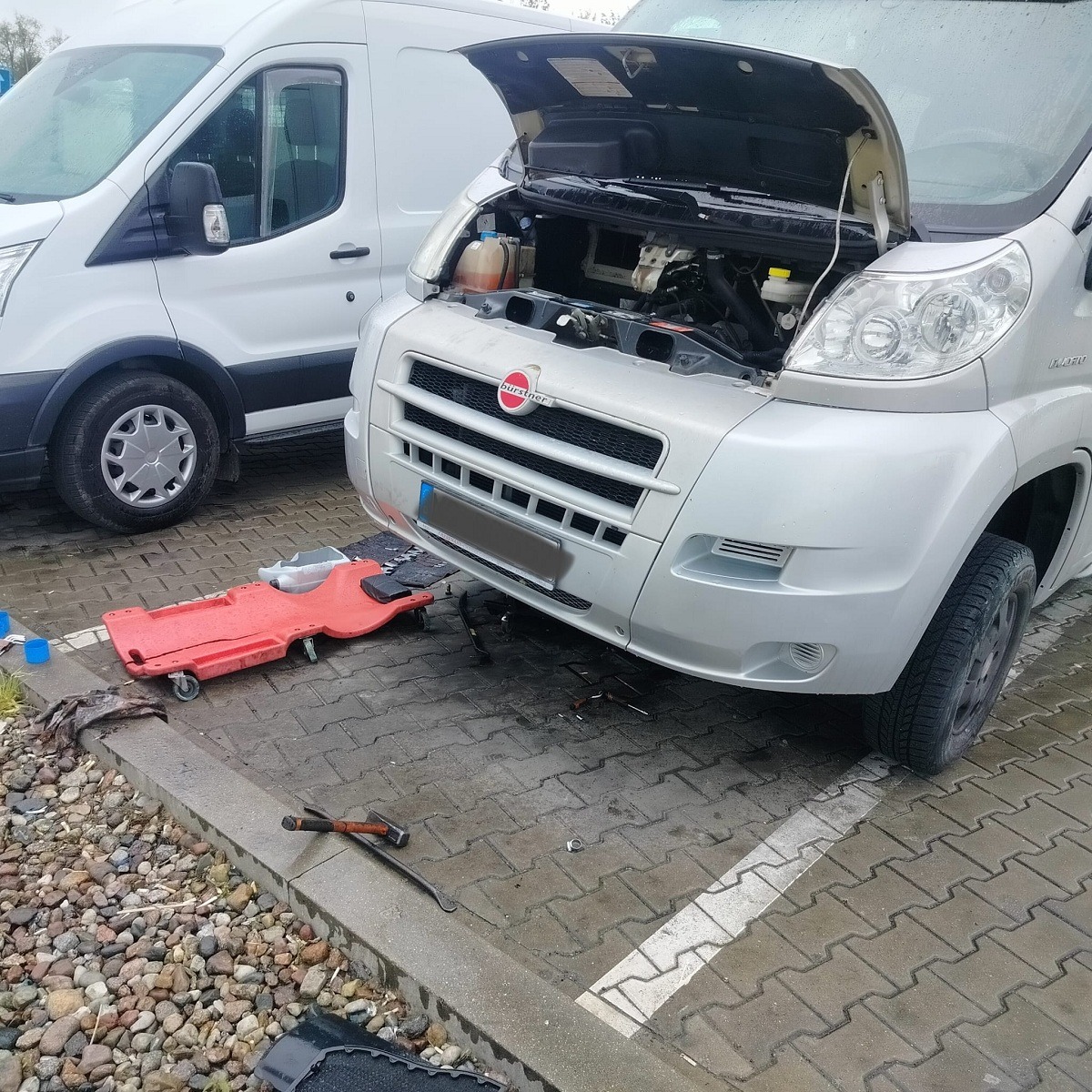 Srebrny bus z otwartą maską, widoczny silnik, obok czerwony leżak warsztatowy i narzędzia na mokrym bruku, drugi biały bus w tle.