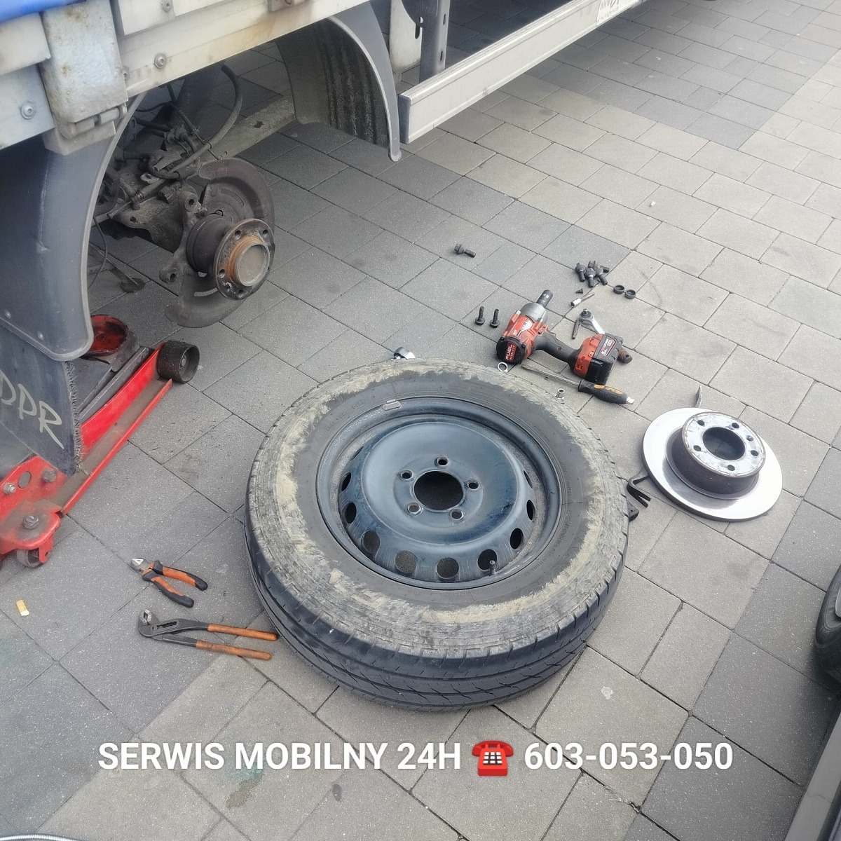 Wymiana koła w pojeździe dostawczym na zewnątrz z użyciem podnośnika, klucza pneumatycznego i nowego koła z tarczą hamulcową. Widoczny numer telefonu serwisu mobilnego.