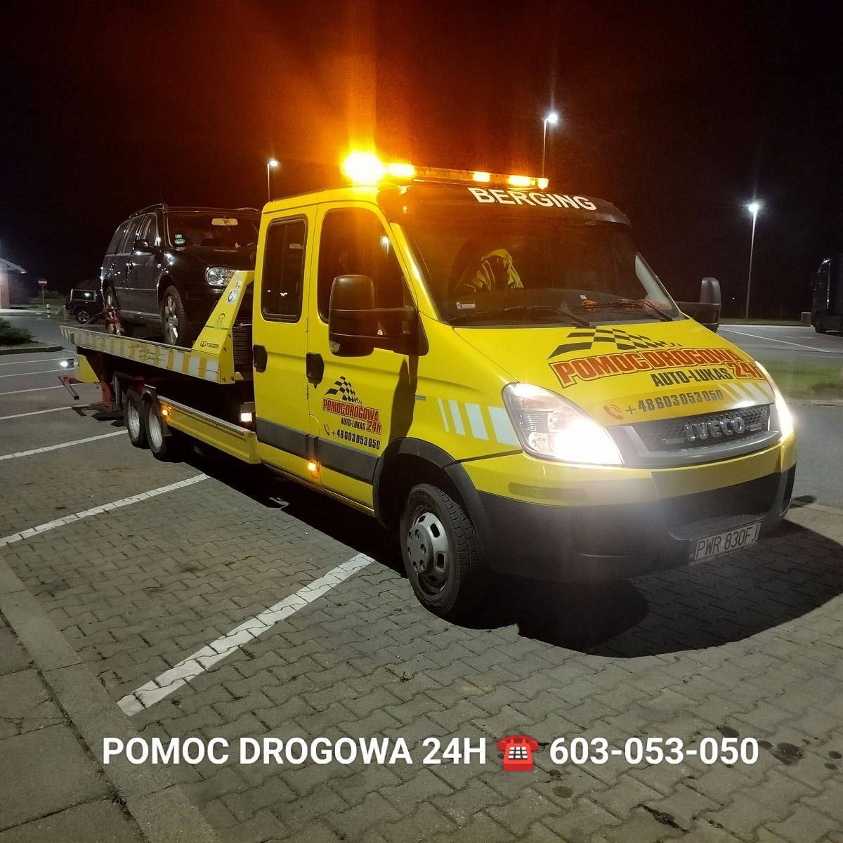 Żółta laweta z włączonymi światłami ostrzegawczymi, przewożąca ciemny samochód osobowy na tle nocnego parkingu z kostki brukowej.