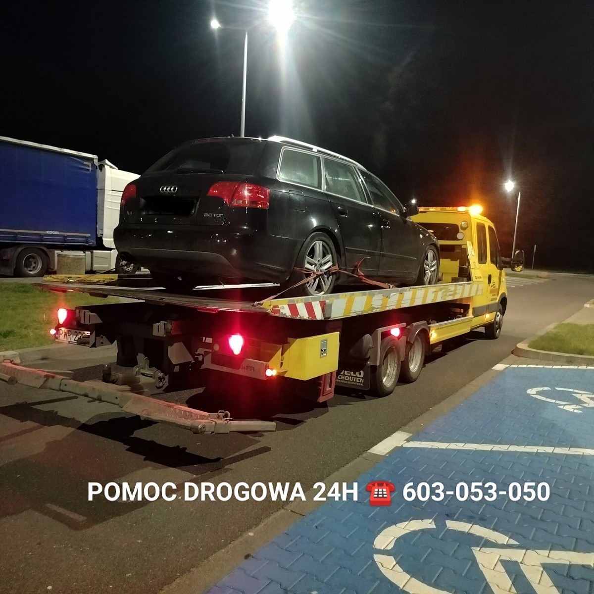 Czarny samochód kombi marki Audi na żółtej lawecie pomocy drogowej z włączonymi światłami ostrzegawczymi, nocą na parkingu.
