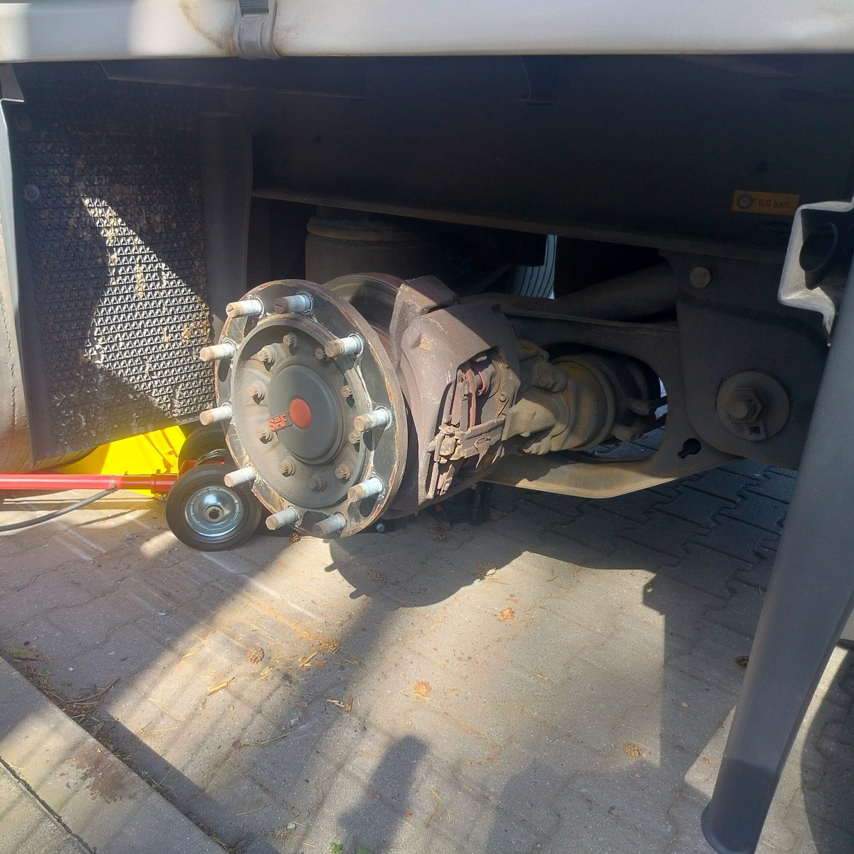 Odkręcone koło ciężarówki z widocznym układem hamulcowym, podparte małym wózkiem transportowym, na brukowanym podłożu.