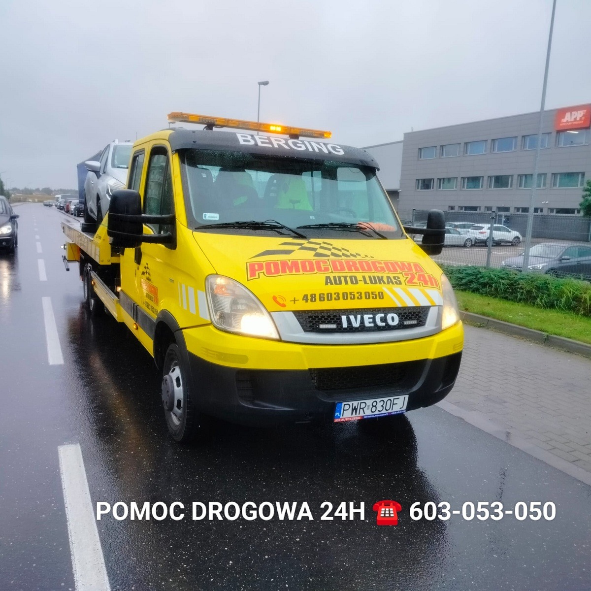 Żółta laweta Iveco z napisem 'Pomoc Drogowa 24h' przewozi srebrny samochód osobowy na mokrej jezdni w pochmurny dzień.