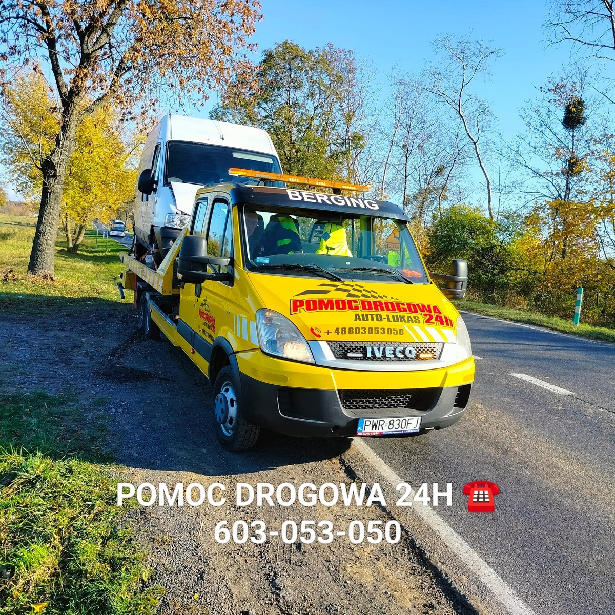 Żółta laweta marki Iveco z napisem 'POMOC DROGOWA AUTO-LUKAS 24h' przewozi białego busa na tle jesiennego krajobrazu.
