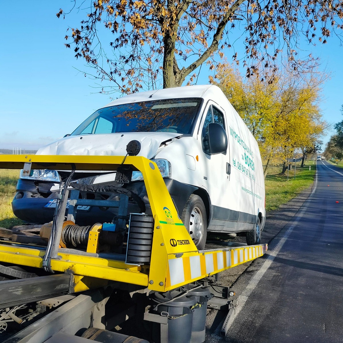 Uszkodzony biały bus z wgniecioną maską załadowany na żółtą platformę holowniczą na poboczu drogi w słoneczny dzień.