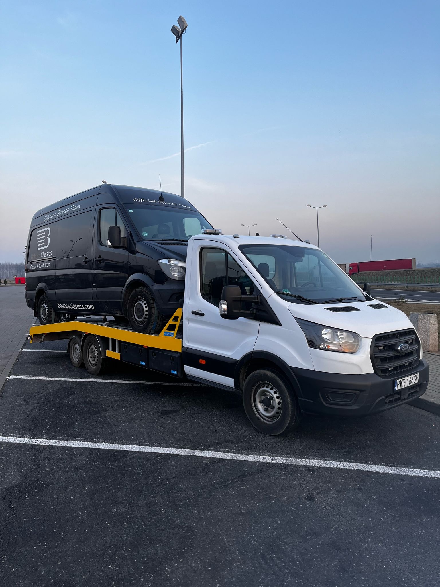 Biała laweta Ford Transit przewozi czarnego busa z napisem 'Belrose Classics' na parkingu w słoneczny dzień.