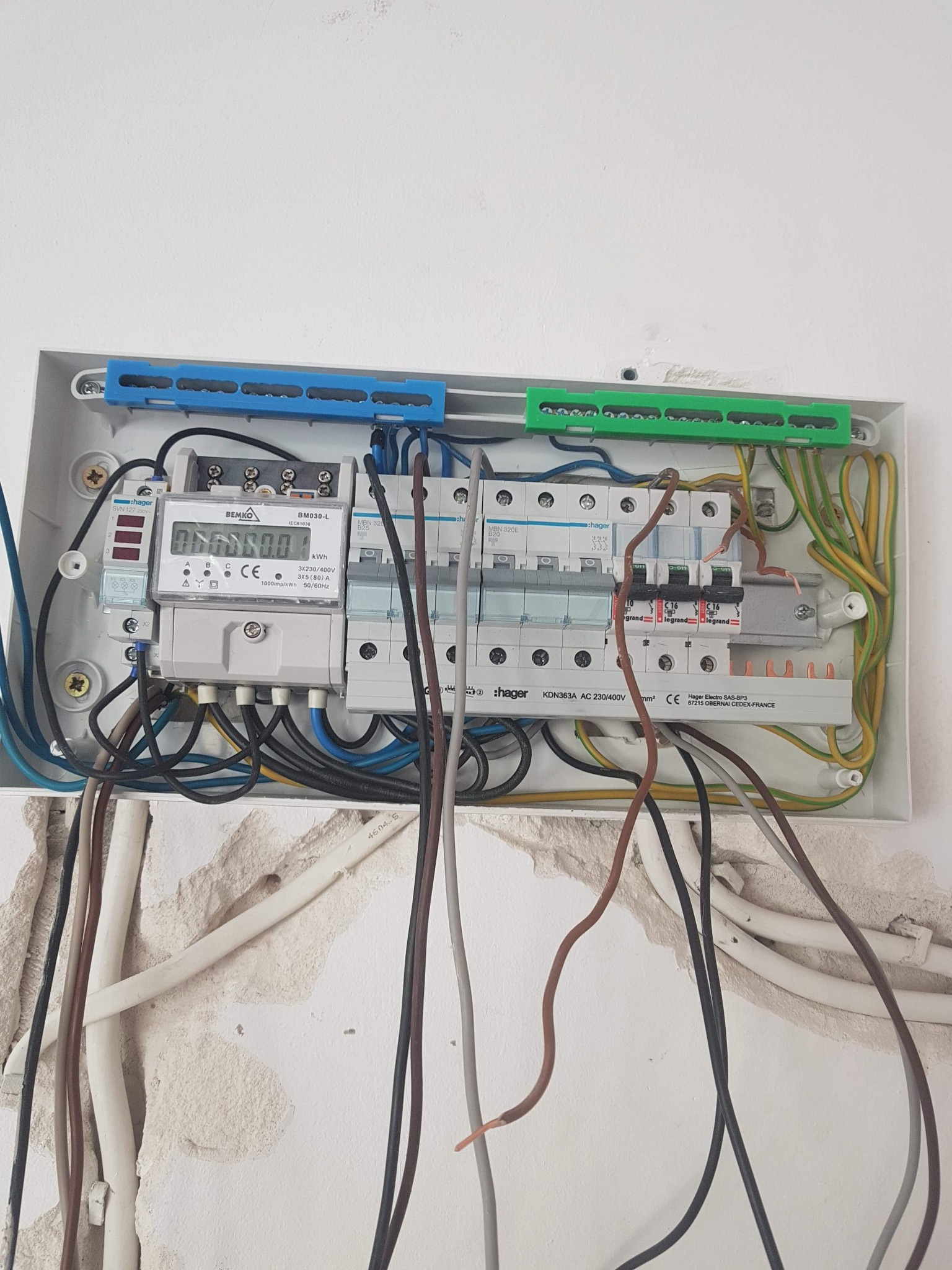 Otwarta skrzynka rozdzielcza z licznikiem energii elektrycznej, widoczne przewody, zabezpieczenia Hager oraz licznik Bemko BM030-L, montaż w trakcie realizacji, ściana z widocznymi ubytkami tynku...