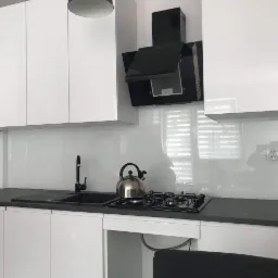 Biała, minimalistyczna kuchnia z czarnym blatem i płytą gazową, na ścianie białe panele szklane, widoczny czajnik na płycie i odbicie fotografa w panelu.