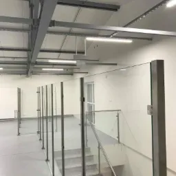 Szklane panele z nierdzewnymi słupkami tworzące zabezpieczenie na antresoli w nowoczesnym wnętrzu przemysłowym, widok na schody i drzwi w tle.