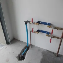 Instalacja wodna z zaworami i rurami miedzianymi, mocowana do ściany na niebieskich uchwytach. Rury wchodzą w wycięcie w betonowej posadzce.