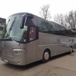 Srebrny autokar marki VOL BOVA z widoczną tablicą rejestracyjną KNS 2625A, zaparkowany na szarym betonowym placu, z drzewami bez liści w tle i odbiciem w szybach.