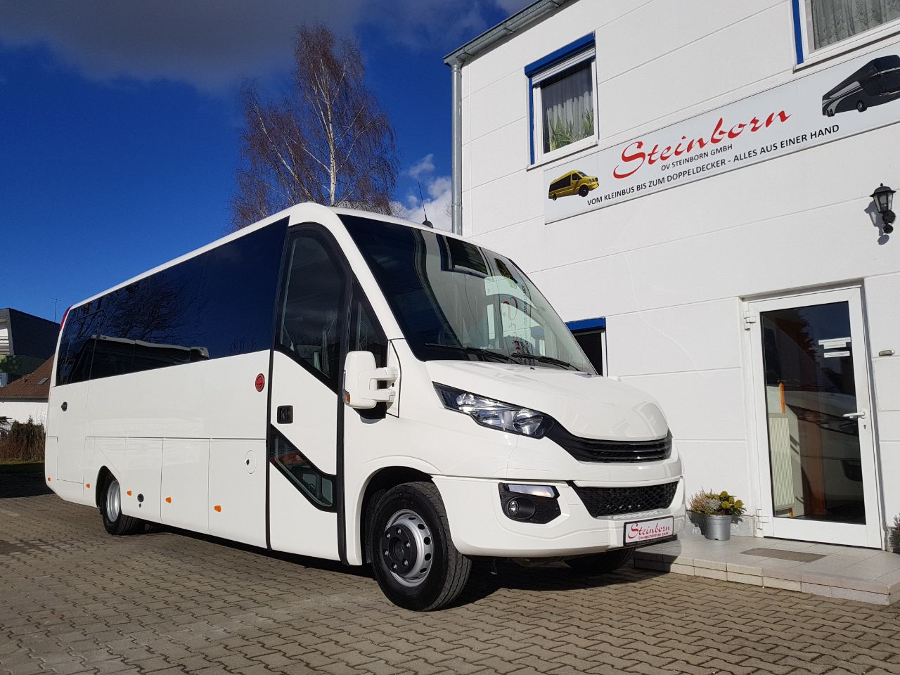 Biały autobus turystyczny Steinborn na tle budynku z logo firmy, błękitne niebo i drzewo w tle.