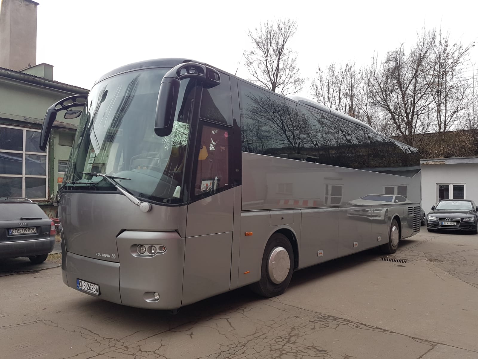 Srebrny autokar marki VOL BOVA z widoczną tablicą rejestracyjną KNS 2625A, zaparkowany na szarym betonowym placu, z drzewami bez liści w tle i odbiciem w szybach.