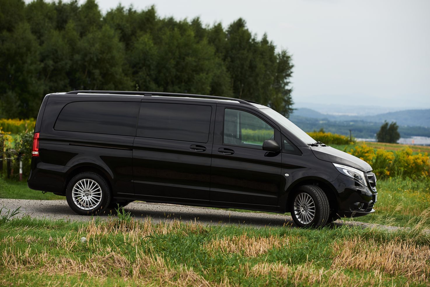 Czarny Mercedes Vito z przyciemnianymi szybami na tle zieleni i zalesionych wzgórz, widok z boku.