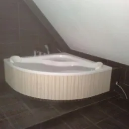 Narożna wanna z hydromasażem, obudowana panelami imitującymi drewno, w łazience wykończonej ciemnymi, kwadratowymi płytkami na podłodze i ścianach pod skosem dachu.