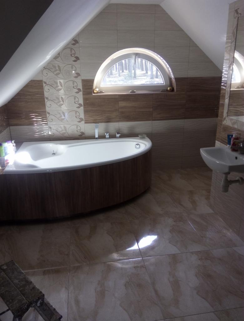 Łazienka na poddaszu z oknem w kształcie półkola, brązowo-beżowe płytki na ścianach i podłodze, wanna z obudową imitującą drewno, umywalka.