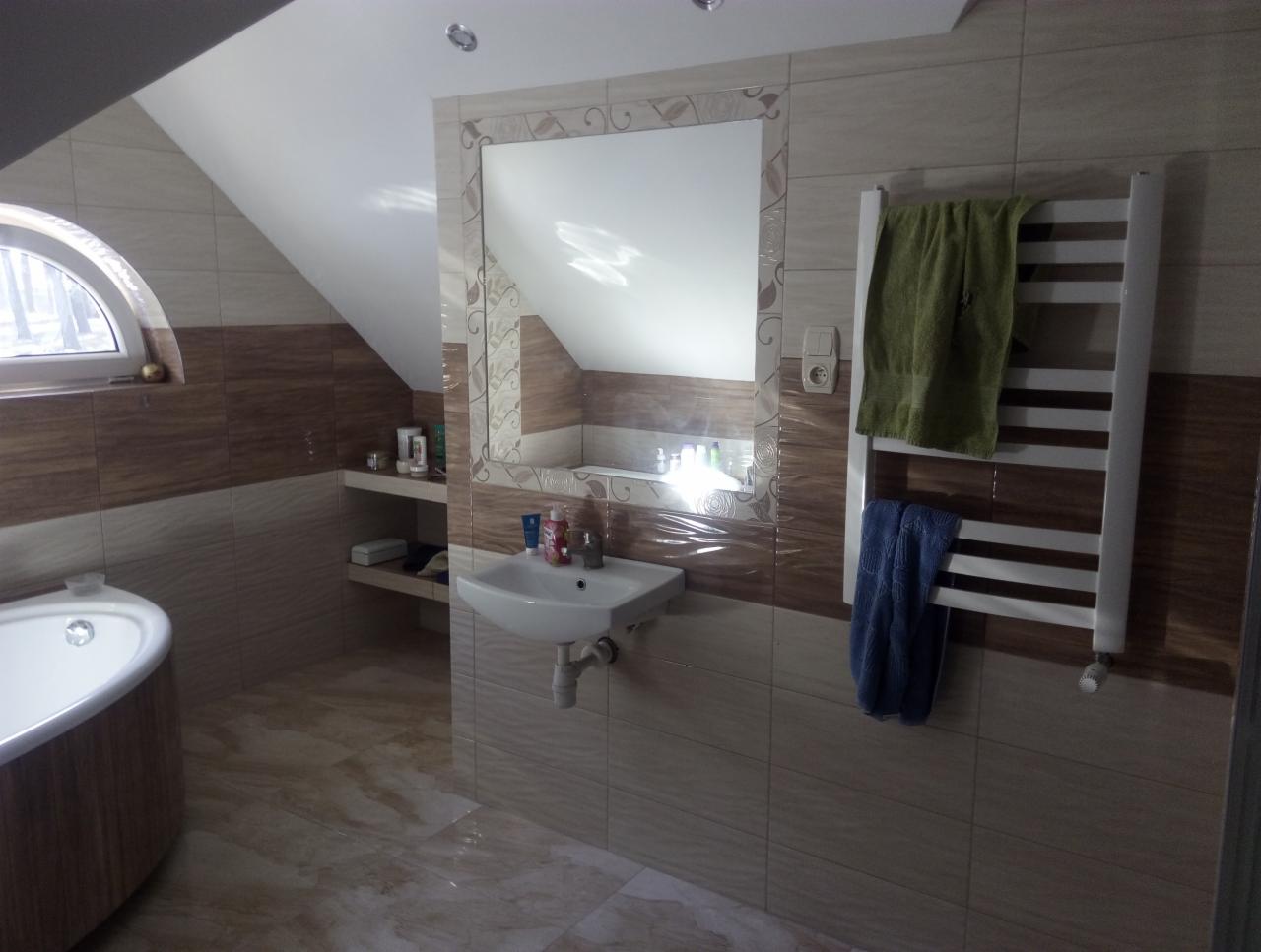 Wykończona łazienka na poddaszu z beżowo-brązowymi płytkami, owalną wanną, umywalką i grzejnikiem drabinkowym z ręcznikami.