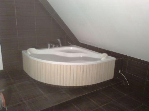 Narożna wanna z hydromasażem, obudowana panelami imitującymi drewno, w łazience wykończonej ciemnymi, kwadratowymi płytkami na podłodze i ścianach pod skosem dachu.