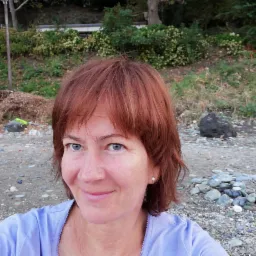 Uśmiechnięta kobieta z rudymi włosami i niebieskimi oczami robiąca selfie na tle plaży z kamieniami i zielenią.