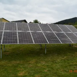 Rząd paneli słonecznych na metalowych wspornikach, widok z niskiej perspektywy na tle domów i zalesionego wzgórza, data 13.05.2020 w prawym dolnym rogu.