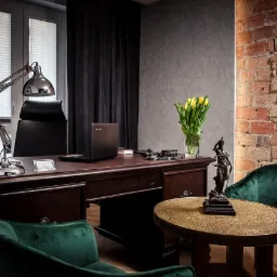 Eleganckie biuro z drewnianym biurkiem, lampą kreślarską, laptopem, bukietem żółtych tulipanów, statuetką Temidy i zielonymi, aksamitnymi fotelami na tle ściany z cegły i szarego tynku