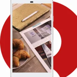 Smartfon wyświetlający zdjęcie otwartej gazety z artykułem o wnętrzach biurowych, obok złoty notatnik, długopis i talerz z croissantami na drewnianym stole.