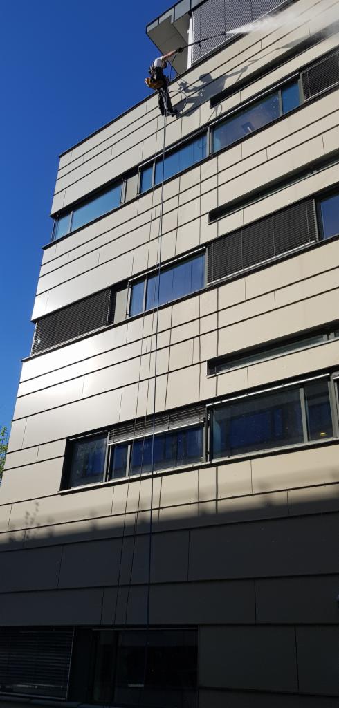 Pracownik na linach myje elewację biurowca myjką ciśnieniową, widoczne liny, okna z żaluzjami i fragment nieba.