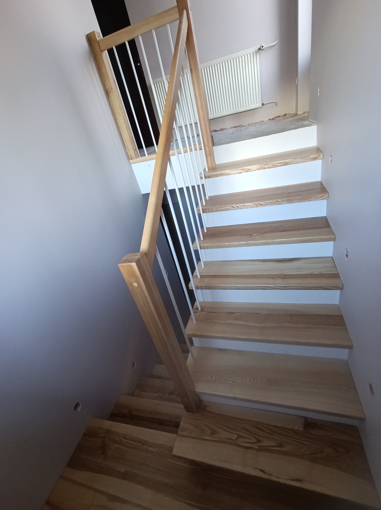 Jasne, drewniane schody z białymi podstopnicami i metalową balustradą, widziane z góry. Ściana w odcieniach szarości, widoczny grzejnik. Ujęcie z perspektywy.