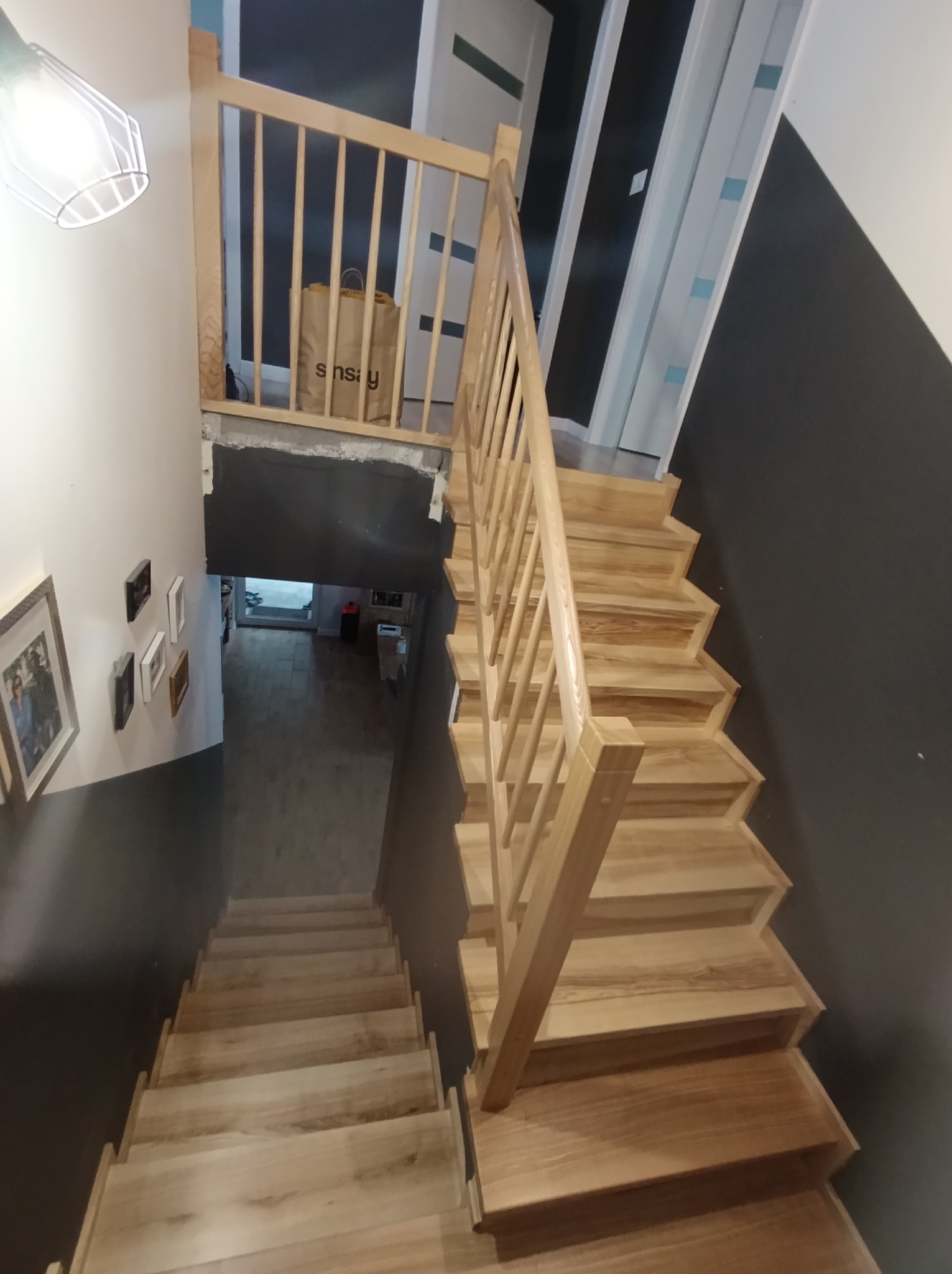 Jasne, drewniane schody z balustradą, widziane z góry. W tle widoczny fragment ściany w kolorze grafitowym i białe drzwi. U dołu schodów widać dalszą część korytarza.