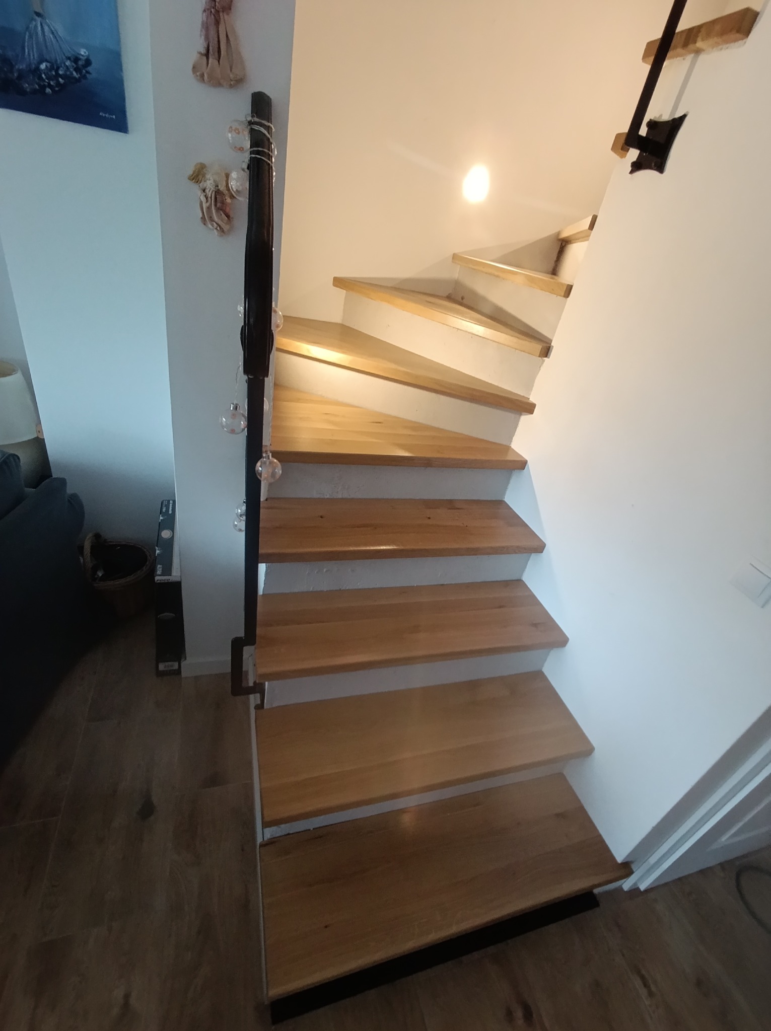 Jasne, drewniane schody z czarną balustradą, oświetlone od góry, prowadzące w górę w białym wnętrzu. Widok z dołu, ukazujący detale wykończenia.