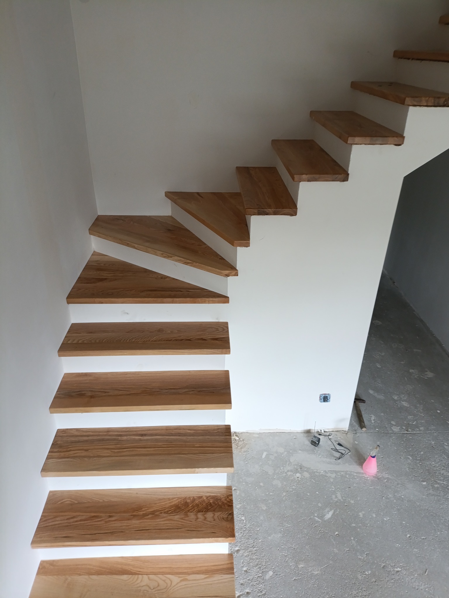 Nowoczesne schody z jasnego drewna, wspornikowe, zamontowane przy białej ścianie. Widoczne detale wykończenia i surowy beton na podłodze.