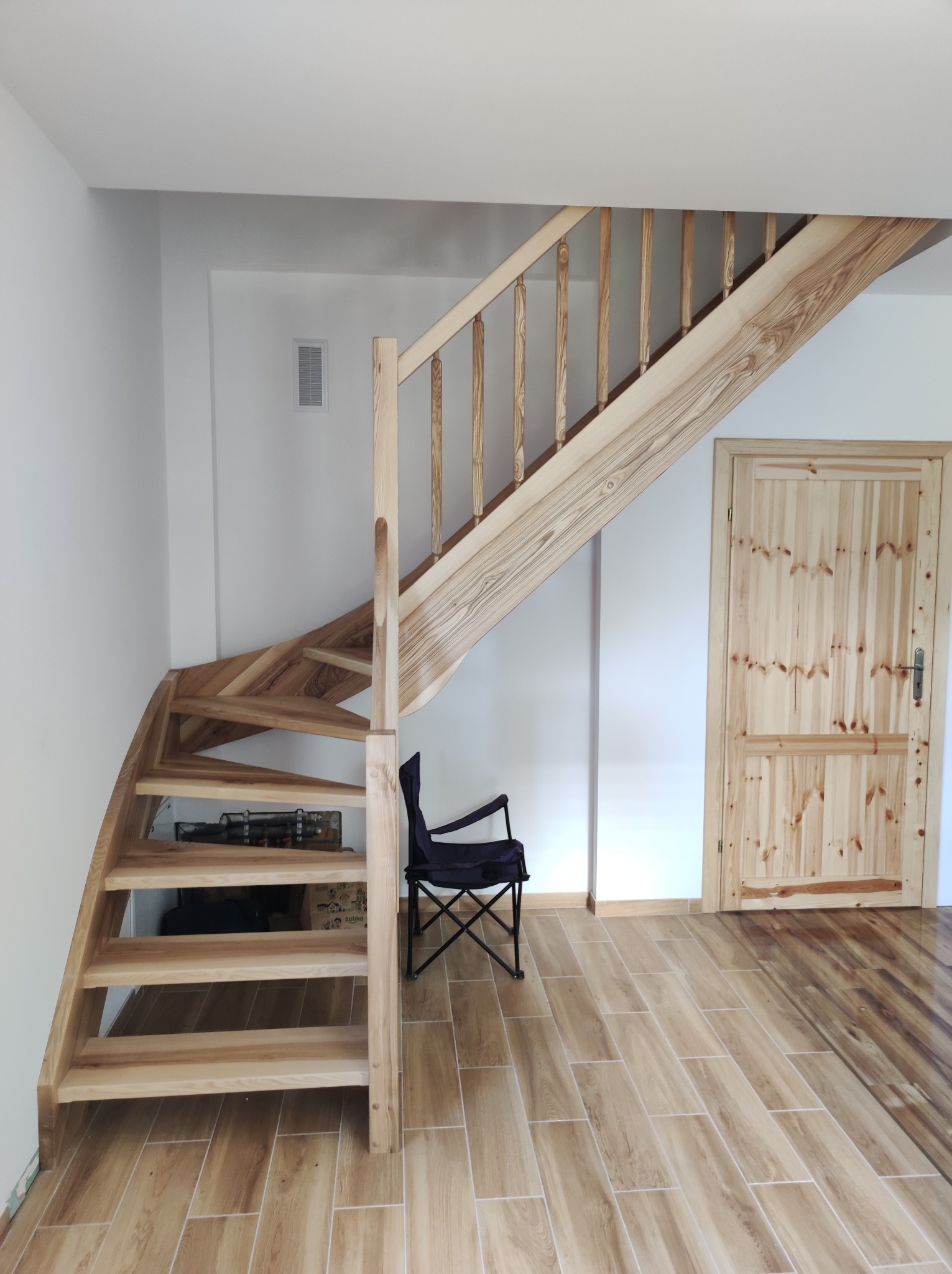 Jasne, drewniane schody kręte z balustradą, prowadzące na antresolę. Pod schodami widoczna przestrzeń do przechowywania. Obok stoi krzesło turystyczne.
