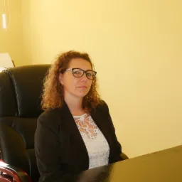 Kobieta w okularach siedzi na czarnym skórzanym fotelu biurowym przed biurkiem, w tle żółta ściana i fragment drukarki.