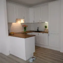 Jasna kuchnia z białymi frontami szafek i drewnianym blatem, oświetlona punktowo, z dekoracjami w postaci kwiatów i małej roślinki doniczkowej, widok z niskiego kąta.