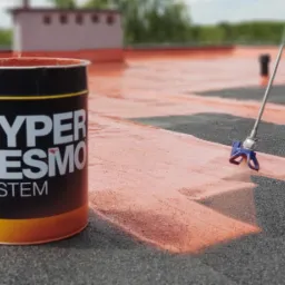 Aplikacja pomarańczowej powłoki Hyperdesmo System na czarny dach za pomocą pistoletu natryskowego, obok stoi puszka z farbą.