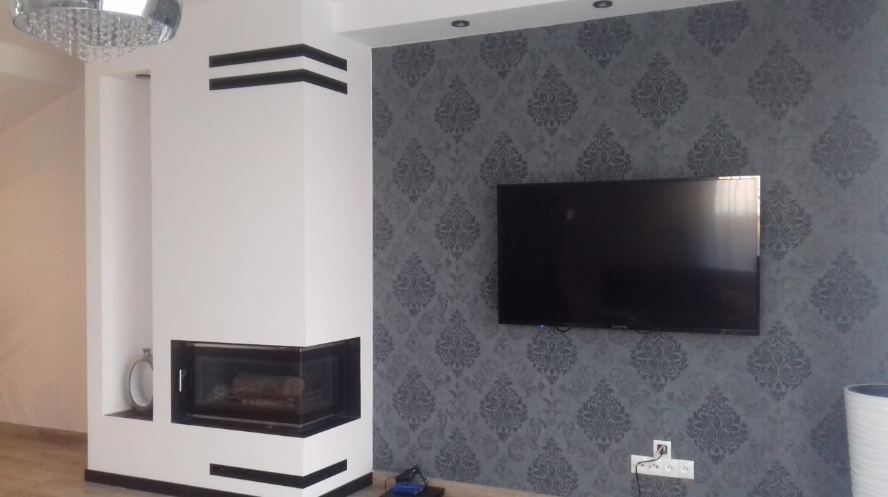 Salon po remoncie: biały kominek z czarnymi detalami, ściana w szare wzorzyste tapety, wiszący telewizor, minimalistyczny design.