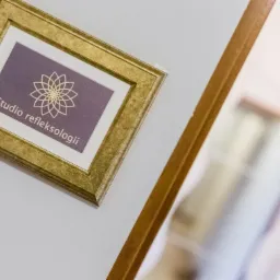 Złota ramka z napisem 'Studio refleksologii' i symbolem kwiatu na fioletowym tle, wisząca na białej ścianie obok drewnianej ramy lustra.