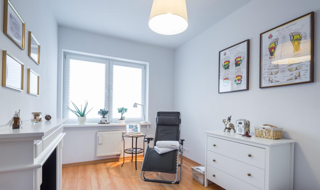 Jasne wnętrze gabinetu z fotelem relaksacyjnym, plakatami z mapami refleksologicznymi na ścianie i dekoracjami na białych meblach.