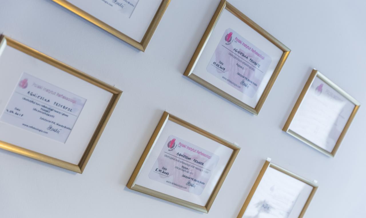 Ściana gabinetu z ramkami zawierającymi certyfikaty ukończenia kursów refleksologii, w złotych ramkach, z nazwiskiem Agnieszka Przybysz.