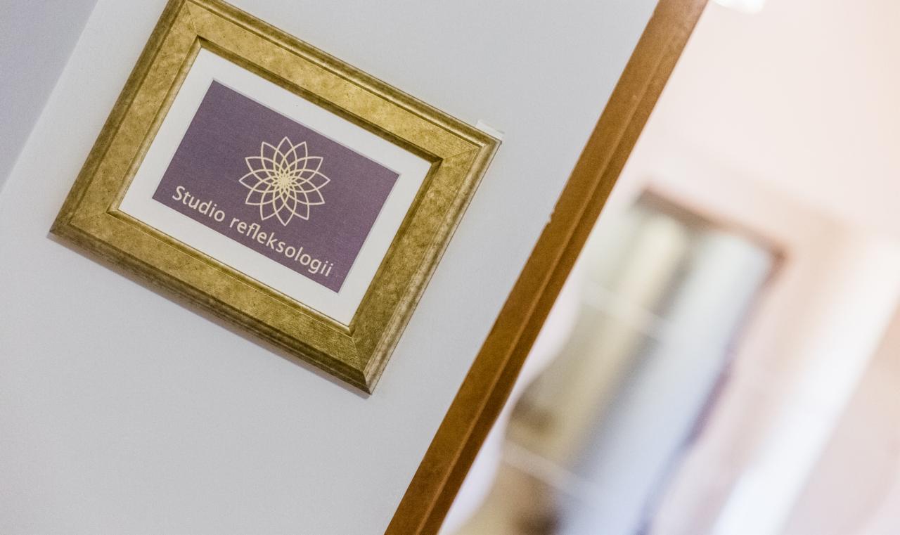 Złota ramka z napisem 'Studio refleksologii' i symbolem kwiatu na fioletowym tle, wisząca na białej ścianie obok drewnianej ramy lustra.