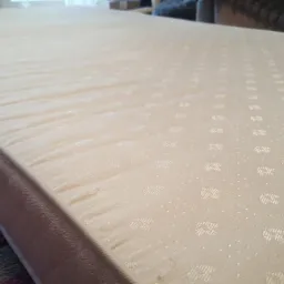 Beżowy materac z delikatnym wzorem, leżący na kolorowym dywanie, sfotografowany z niskiego kąta ukazującego jego grubość.