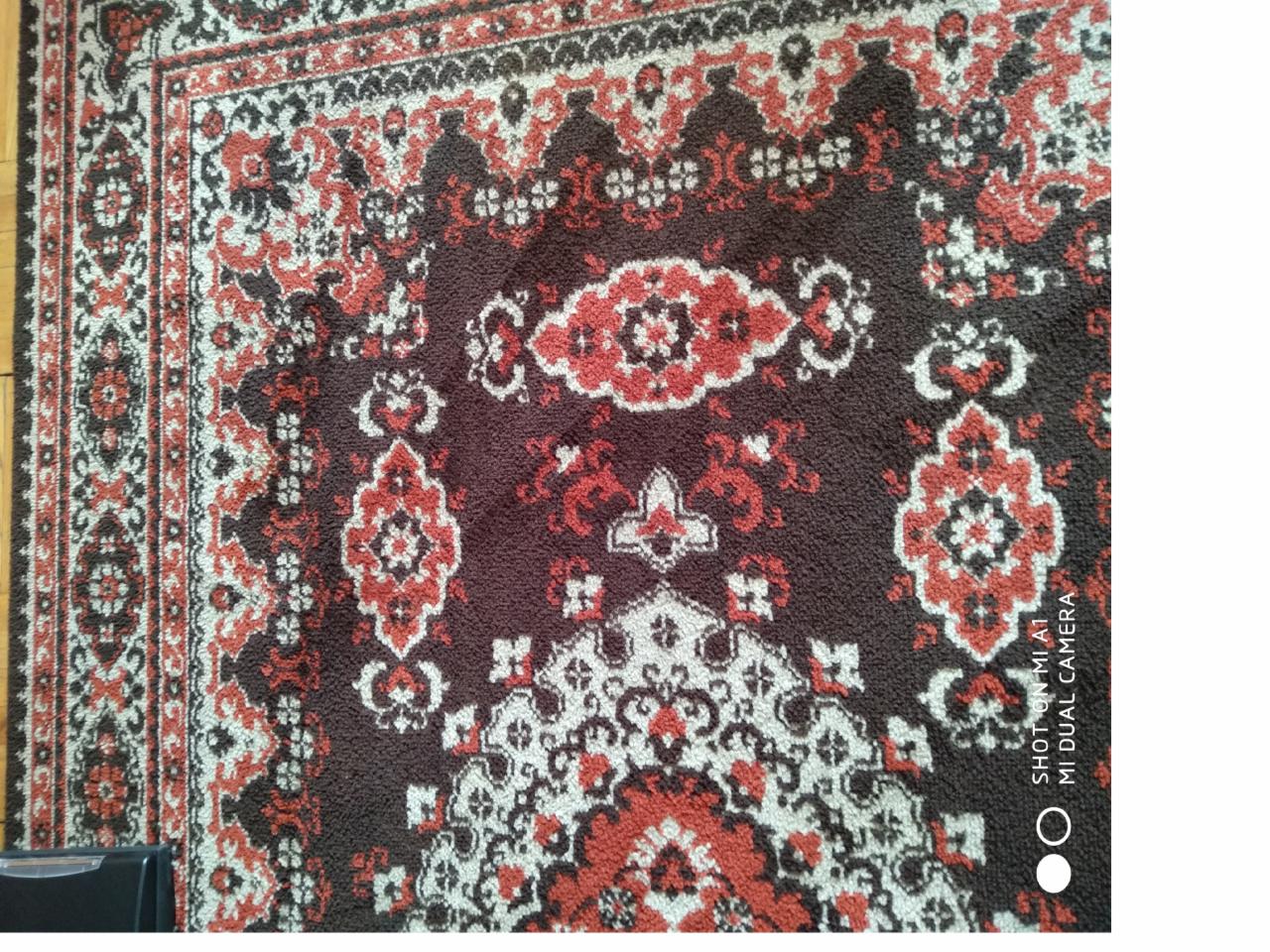 Fragment wzorzystego dywanu z dominującymi kolorami brązu, czerwieni i bieli, sfotografowany z bliska, z widocznym logo aparatu w prawym dolnym rogu.