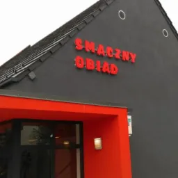 Zewnętrzny widok budynku z napisem 'SMACZNY OBIAD' w czerwonych literach na szarej ścianie, czerwony daszek nad wejściem z przeszklonymi drzwiami.